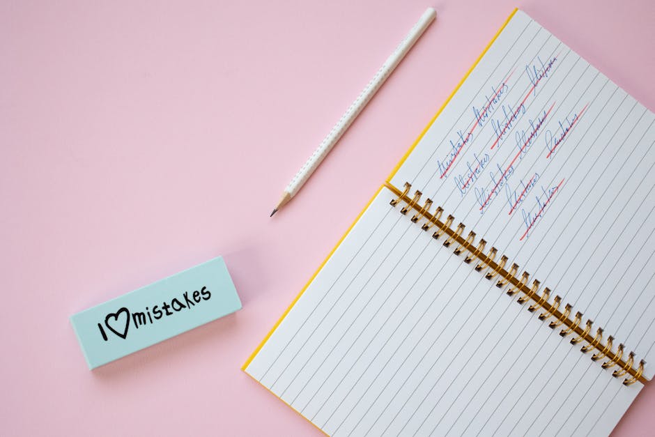 Flat lay of a spiral notebook and eraser on a pastel pink background with crossed out words.